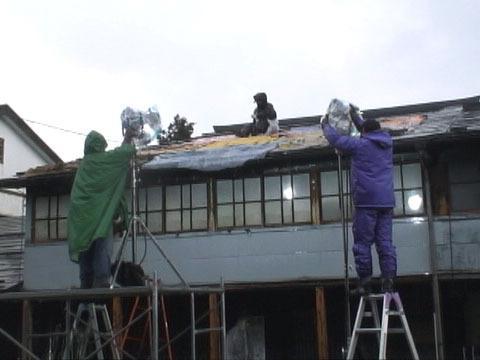 Tournage dans le vieux village aux maisons de bois. Le mauvais temps s’est invité. Aujourd’hui on tourne une scène d’intérieur dans la boutique du teinturier,
mais l’équipe technique s’active de l’extérieur. Montés sur des échelles, les éclairagistes installent de gros spots sur pied qu’ils dirigent vers les fenêtres pour éclairer à travers les vitres l’intérieur de la boutique comme par une belle journée de soleil. Sur le toit de vieilles tôles, la pluie produit en s’écrasant des toc, toc, ploc, ploc, qui couvrent les voix des comédiens.
Deux décorateurs montent sur le toit pour le couvrir de vieilles couvertures et atténuer lebruit des gouttes d’eau. Toute l’équipe en extérieur travaille sous l’averse, gestes précis et volontaires dans les ponchos imperméables, silhouettes de couleurs dans le village noir de bois trempé.