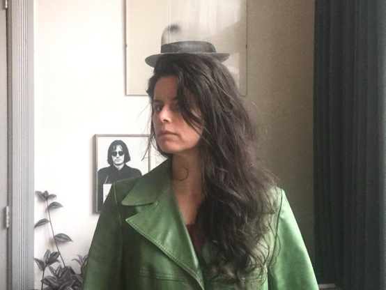 Standing in front of a framed photo of a hat (an artworklby Joseph Beuys) it looks like Gemma is wearing it. Long dark wavy hear comming from underneath it, the old schooljhat goes well with the green leather transh-coat.