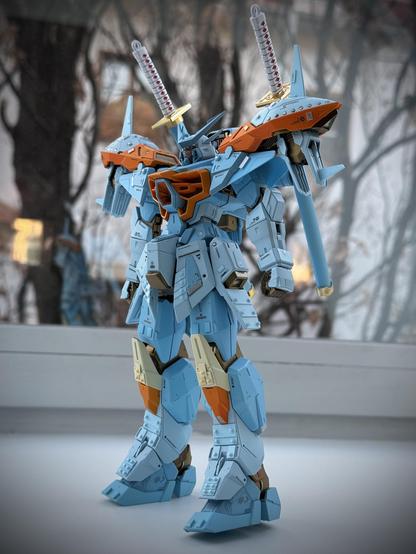 A detailed model of a mech, predominantly in blue and orange colors, featuring katana-like weapons on its shoulders. The background includes soft, blurred tree silhouettes. The figure is posed against a light surface, showcasing its mechanical design.
