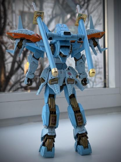 The image features a detailed model of a blue robotic figure with orange accents, striking a dynamic pose. It has two large blue weapons resembling swords and is set against a window with blurred tree branches in the background.