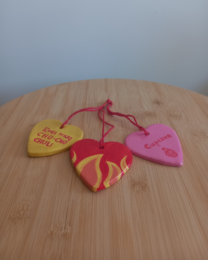 Foto en la que aparecen 3 colgantes en forma de corazón con un hilo rojo de yute anudado cada uno sobre una mesilla de madera. El de la izquierda es amarillo y tiene escrito en rojo "Eres muy CHU-CHU-CHULI", el del medio es rojo con llamas en su parte inferior y el de la derecha es rosa con la palabra "Cupcake (pastelito)" escrita en rojo y un pastelito con un sombrero de guinda dibujado en rojo.