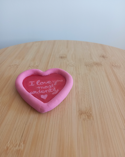 Foto de un portavelas en forma de corazón sobre una mesilla de madera. El interior está pintado en rojo, el texto en el centro del objeto y el borde está pintado en rosa. Se lee: "I love you, most ardently (La amo, ardientemente)" rematado en un corazón.