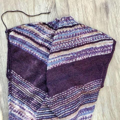 A flat lay view of a tee being knit in purples, it's shown on a light wooden background. One armhole is finished with a simple knit bind-off, the other with an I-cord finishing.