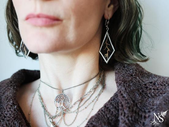 A woman's neckline with the bottom of her face. We see earrings in the shape of angular drops, made of glass and set in a silvery contour, containing a real piece of roots. At her neck, a multi-chain necklace with a choker of a tree of life pendant.
