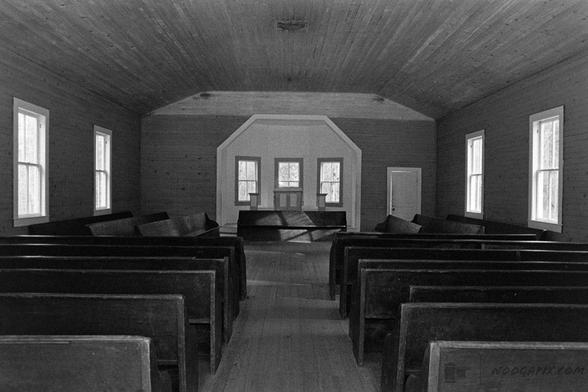 A black-and-white photo of a simple wooden church interior, featuring rows of wooden pews and a small pulpit lit by natural light from the windows.