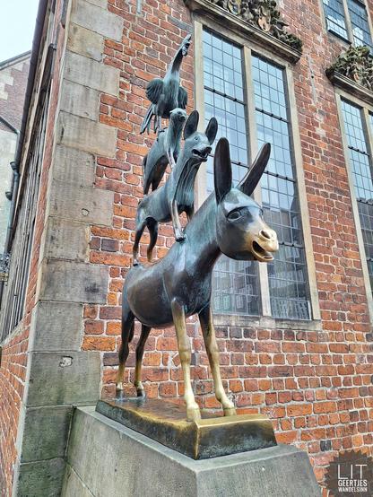 Auf einem Esel steht ein Hund. Auf dem Hund eine Katze. Auf der Katze ein Hahn. Die Skulptur steht vor einem Backsteinhaus.