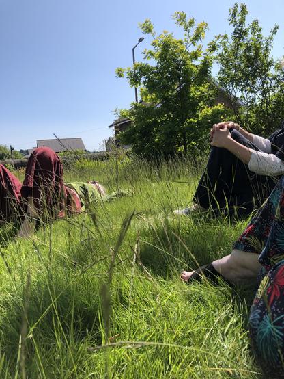 A photo of Vicky lying on the grass and you can also see the legs of Trudy and Rah. There are some trees in the background and houses and clear blue sky