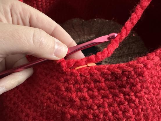 A close-up of a person's hand holding a pink crochet hook, working on a red crochet item.  A peach colored stitch marker indicates progress is at the beginning of a new row.  