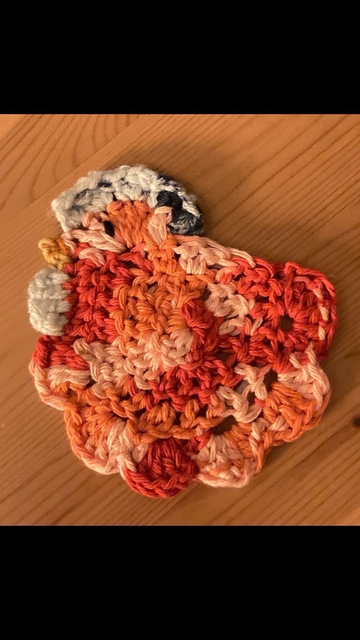 Photo ID: image of an orange and light orange-y yellow white and blue crocheted chicken coaster. 
