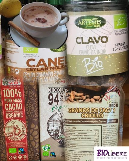 Imagen de varios productos alimenticios orgánicos: una taza de bebida caliente con especias, un bote de clavos, una bolsa de canela molida en polvo, una barra de chocolate negro 100% cacao de República Dominicana y una bolsa de granos de cacao criollo. Se aprecian detalles como el etiquetado que indica el origen y la certificación orgánica de los productos.