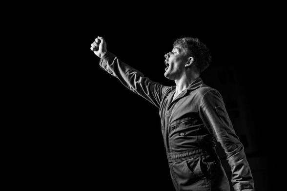 A man in a grey jumpsuit raises his fist passionately while shouting, set against a dark background.