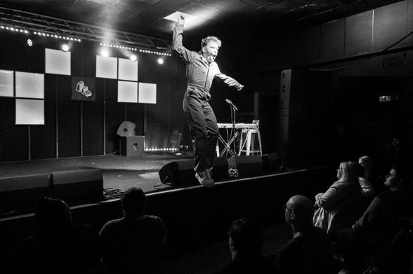 A performer in a jumpsuit is energetically dancing on stage, with a microphone and keyboard setup in the background. The audience, seated in the foreground, is watching attentively. The scene is in black and white with stage lighting creating a dramatic atmosphere.