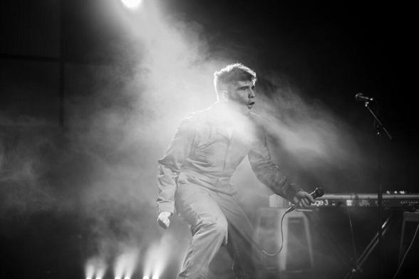 A performer in a jumpsuit passionately sings into a microphone on a stage filled with smoke, with dramatic lighting illuminating from above.