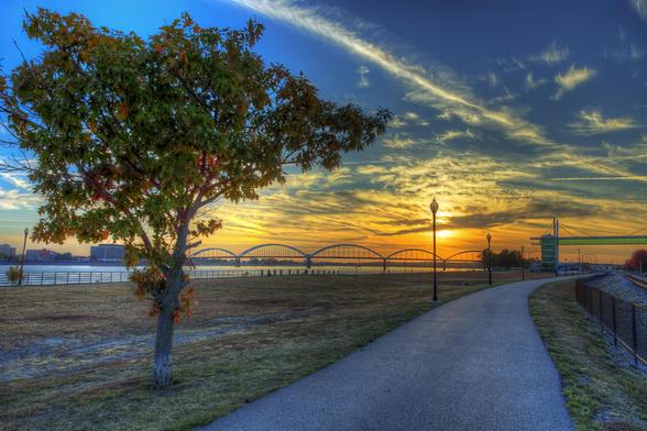 A vibrant sunset casts warm tones across a serene Davenport riverside path, with a solitary tree and arching bridge enhancing the tranquil scene.