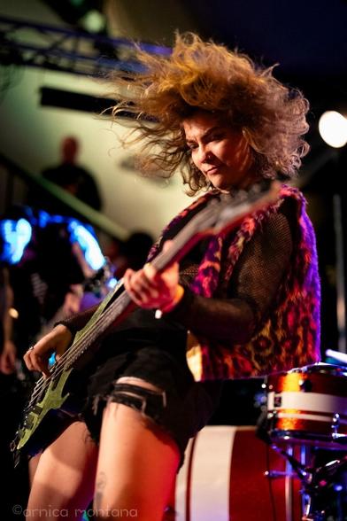 A musician playing bass guitar on stage, with hair flying and a vibrant outfit. The performance captures an energetic moment, with a blurred background showing audience members and instruments.