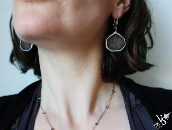 A woman's neckline with the bottom of her face. We see earrings with a black background and skeleton hydrangea petals on them. They are made of glass and set in a silvery contour and the shape is the same as the petal.
