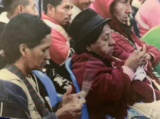 A group of Indigenous women and men are seated in blue chairs at a gathering. Two persons in the foreground, dressed in traditional clothing, are weaving or crafting with their hands. One person wears a black hat and a red shawl, while the other wears a woven scarf. The background shows more people in colorful clothing, engaged in the event.

Photo courtesy of Luisa Bagope