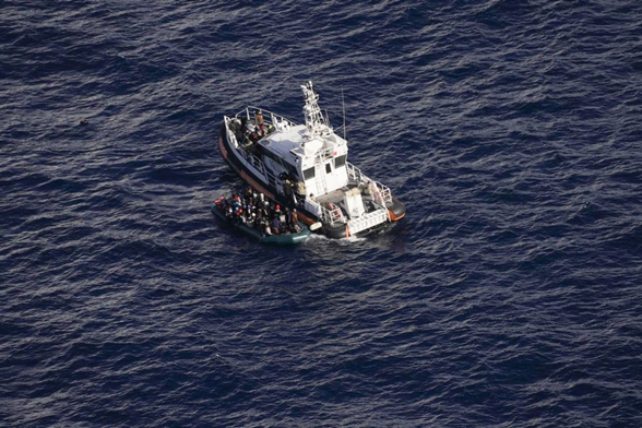 Ein Küstenwachboot (vom Typ her eines der letzten modernen von Italien an Libyen gespendeten Patrouillenboote) liegt mitten auf dem Meer längsseits eines überfüllten Schlauchboots.