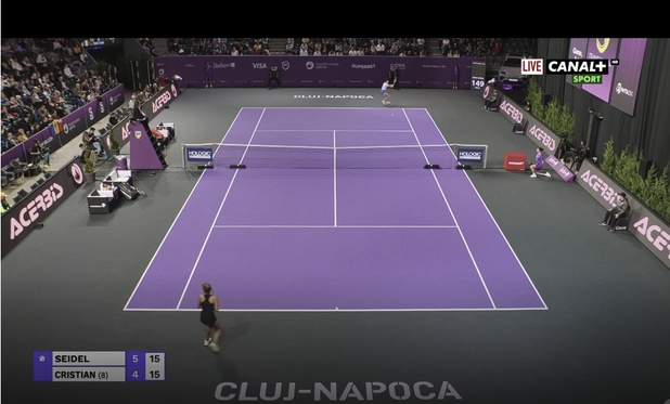 Ella Seidel (far court) plays Jaqueline Cristian, at the WTA 2025 Transylvania Open, Day 2, Round of 32 play on Center Court, at the BT Arena in Cluj-Napoca, Romania. The court surface color is light-purple white lines; the outer court surface is gray-green. Walls behind players are purple. Medium-height black digital displays with white graphics: run parallel to the length of the court, displaying repeating brand promotions. Text "Cluj-Napoca" appears behind each baseline in large white block letters. The stands appear full of spectators. the players seats are at left near the net, on either side of the elevated chair umpire (her tall chair has purple sides with a Banca Transilvania shield logo).

The on-screen graphic says "Live Canal+ Sport" in the upper-right. The score at bottom-left shows Seidel leading the 1st set 5-4 at 15-15.

Court Branding & Signage: 
Net signs: Holologic (near the posts; rectangular & dark-purple with white lettering)
Digital signs running parallel to the court: Acerbis
Walls behind players (white logos): Banca Transilvania, Nova Power and Gas, Superbet, Visa, Porsche, Radisson Blu Cluj, Cavit, Evergent Investments, Hexagon, Regina Maria, Rivus, etc. 