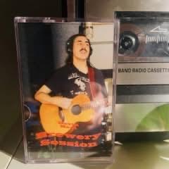 Eine Kassette mit einem Mann mit Schnurrbart, der eine Akustikgitarre spielt. Die Kassette trägt die Bezeichnung "Discovery Session" und befindet sich neben einem Kassettenspieler.