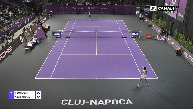 Viktoriya Tomova (far court) plays Olga Danilović, at the WTA 2025 Transylvania Open, Day 2, Round of 16 play on Center Court, at the BT Arena in Cluj-Napoca, Romania. The court surface color is light-purple white lines; the outer court surface is gray-green. Walls behind players are purple. Medium-height black digital displays with white graphics: run parallel to the length of the court, displaying repeating brand promotions. Text "Cluj-Napoca" appears behind each baseline in large white block letters. The stands appear partially full of spectators (some of the rows of chairs near the court are empty). the players seats are at left near the net, on either side of the elevated chair umpire (her tall chair has purple sides with a Banca Transilvania shield logo).

The on-screen graphic says "Live Canal+ Sport" in the upper-right. The score at bottom-left shows the score in the 1st game of the 1st set, tied at 30-30.

Court Branding & Signage: 
Net signs: Holologic (near the posts; rectangular & dark-purple with white lettering)
Digital signs running parallel to the court: Acerbis
Walls behind players (white logos): Banca Transilvania, Nova Power and Gas, Superbet, Visa, Porsche, Radisson Blu Cluj, Cavit, Evergent Investments, Hexagon, Regina Maria, Rivus, etc. 