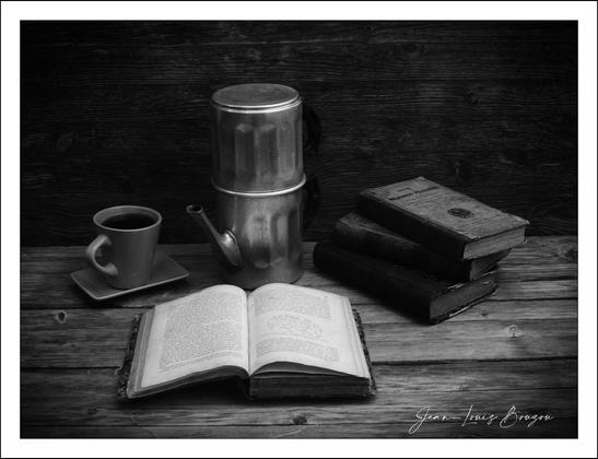 
The image presents a serene still life, evoking a sense of quiet contemplation. At the center lies an open book, its pages slightly fanned, suggesting an invitation to delve into its contents. The book is well-worn, hinting at stories well-loved and wisdom shared.

To its left, a sleek metallic coffee pot glimmers softly, accompanied by a delicate cup of coffee placed on a small saucer. 

Nestled beside the book, a small stack of similarly aged books stands tall, echoing a legacy of knowledge. The wooden surface beneath them further enriches the composition, creating a rustic feel that enhances the overall atmosphere of tranquility and nostalgia.

The black-and-white palette intensifies these elements, focusing attention on shapes and textures rather than color. This choice adds a layer of timelessness, inviting viewers to ponder the simple yet profound pleasures of reading, learning, and enjoying a warm beverage, perhaps hinting at a pause from the hustle of modern life.
