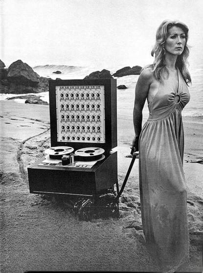 Woman wearing a dress walks along a beach dragging a trolley holding a multitrack reel-to-reel tape recorder.