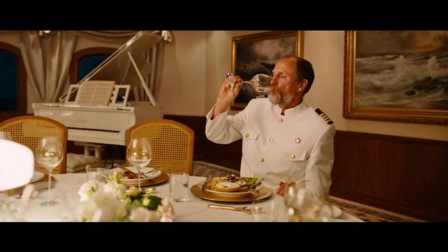 A distinguished man in a white captain's uniform is sitting at a dining table, sipping from a wine glass. The setting features a luxurious interior with a grand piano in the background and elegantly set plates and glasses on the table.