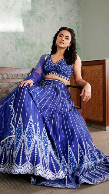 Shraddha Srinath enchants in a blue lehenga by Chamee and Palak, featuring intricate white thread embroidery and a plunging V-neckline. The ensemble is paired with a flowing chiffon dupatta, adding a touch of grace. Captured by Shivasai at 'Sambhrama' and styled by Nidhi Jeswani, Shraddha's look is completed with bold eyeliner, glossy red lips, and defined brows by makeup artist Divya Sunder Rajan, along with elegant 'Mon Tresor' jewellery pieces.