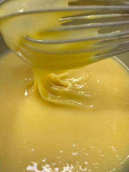 Hellgelbe Creme (Eierlikörfüllung für Pralinen) in einer Metallschüssel. Ein Schneebesen der in die Creme getunkt wurde und jetzt wieder über der Creme ist, zieht einen Teil der Creme nach oben.