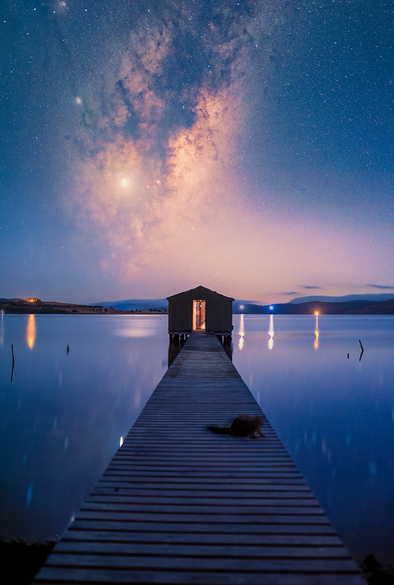 The scene is set at the terminus of a small wooden dock and a boat shed on a shallow, calm bay. The boat shed is illuminated from the interior through an open door. Above the boat shed is the Milky Way rising with Jupiter embedded near Scorpius. There is several distant lights across the bay, including a castle to the scene left. These lights and the Milky Way are reflected in the bay’s calm water. The first traces of sunlight are starting to illuminate the sky, and contaminating the rich blues of the scene with warm tones. On the dock, there is the ghostly apparition of a Brushtail possum briefly trapped in the scene. 