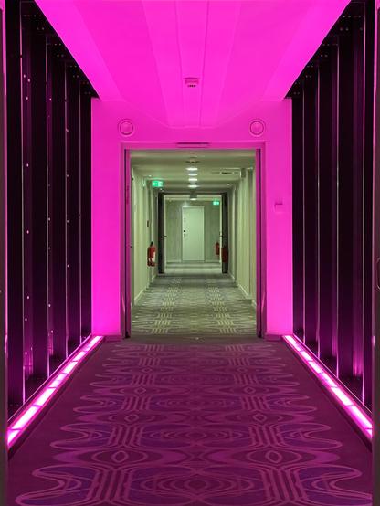 Langer Hotelgang. Am Ende eine weisse Tür. Der Gang hinten ist in grünes, vorne in pinkes Licht gehüllt.
