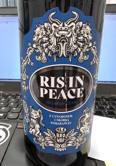 Butelka piwa „Ris in Peace” z Ale Browaru stoi na klawiaturze laptopa. Po środku etykiety medalion z nazwą i słabo widocznym granatowym napisem „Winter 2018”, poniżej napis „z cynamonem i skórką pomarańczy”. Dookoła na granatowym tle czarno-białe rysunki smoków w stylu… hmm komiksowo azjatyckim? Ale są też czaszki które trochę przywodzą mi na myśl sztukę Indian z południowej ameryki. Na dole logo browaru, szklanka w stylu nonic z wpisanym słowem AleBrowar pociętym na trzy literowe sylaby. 