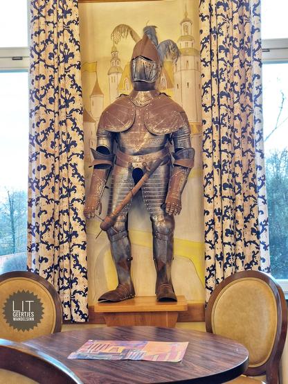Ein rostiger Ritter mit Morgenstern zwischen zwei Fenstern. An der Wand im Hintergrund sind Zeichnungen von Gebäuden zu erkennen. Vor dem Ritter steht ein Tisch und Stühle.