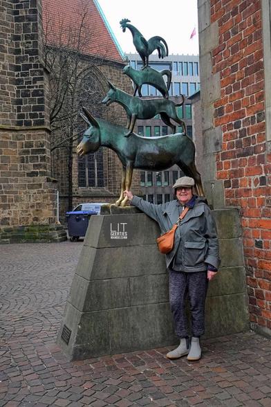 Eine Frau mit Schirmmütze und einer Kängurutasche hält ihre rechte Hand an das linke Bein eines Esels. Darüber stehen ein Hund, eine Katze und ein Hahn. Die Skulptur ist grün, die Schnauze des Esels und seine Beine zeigen blankes Metall.