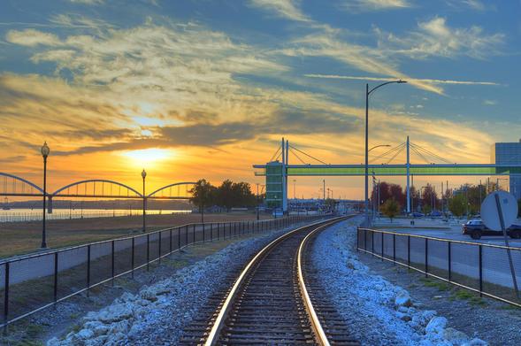 Railroad tracks curve toward a vibrant sunset, with the Centennial Bridge and expansive sky creating a picturesque scene. 