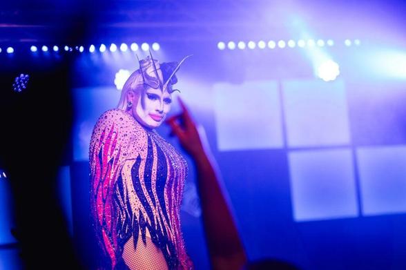 A performer in a vibrant, glittering costume with spikes and dramatic makeup poses confidently on stage, surrounded by purple lighting and a cheering crowd.