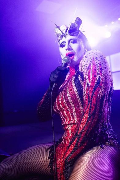 A performer in a glittering red and black costume with fishnet stockings, holding a microphone and striking a pose, under vibrant purple lighting.