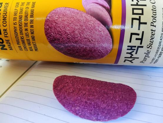 A crisp (UK) or chip (US) shaped like a Pringle crisp, a formed saddle-shape or hyperbolic paraboloid. It is a light purple colour and is in front of the cardboard tube it came in. The tube is yellow and white, and has a photo of the crisps on, with English and Korean text. 