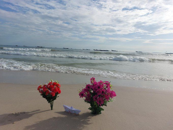 Zwei Blumensträuße, leuchtend rot und pink, stecken im Sand an einem Strand. Zwischen ihnen steht ein Papierschiffchen. Wellen branden im Hintergrund ans Ufer.