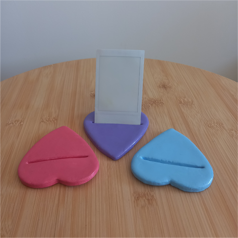 Foto a tres portafotos planos en forma de corazón sobre una mesilla de madera. El de la izquierda es rosa, el del medio es morado y en él hay una foto Polaroid blanca vertical, y el de la derecha es azul.