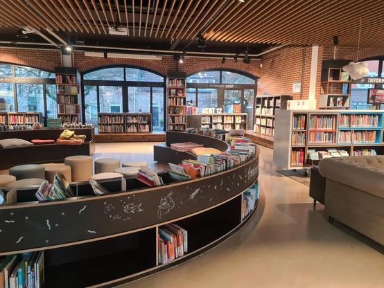 A view into the library with a round low bookshelf around a seating area and rectangular shelves full of books around the edges. The corner of a cream sofa is just visible on the right Ns there are floor to ceiling arched windows around the room. White tile(?) Floor and brick walls.