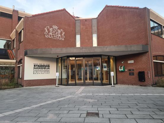 The outside of the library. Light grey tile path leading to a brick building with a large glass entryway. The building has a white section of wall on the left with black letters that say "Stadhuis Harderwijk" and some other smaller text. There is also a white crest on the bricks with a crown and some lions.