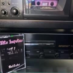 Eine Kassette mit der Aufschrift "Nite Impetus - Solitude" befindet sich vor einem Kassettenspieler, mit einem Vintage-Tape-Deck im Hintergrund.