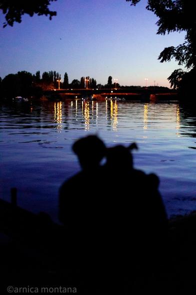 A silhouette of a couple sitting by a calm river at dusk, with reflections of lights on the water and a bridge in the background. The sky transitions from blue to purple.