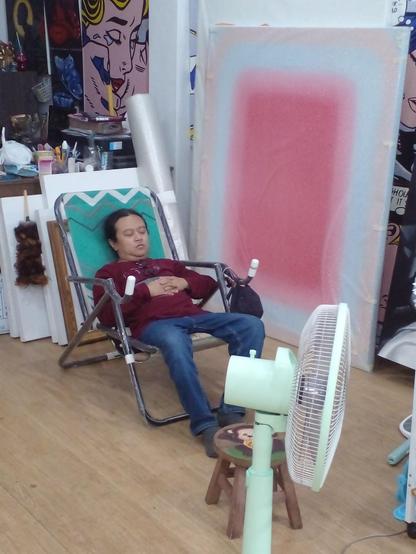 a photo of geng sleeping in a camping chair, a fan in front of him, paintings around him, his hands folded over his chest 