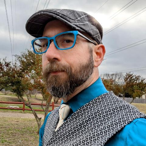 A photo of me, a 46 year old white man with a shaved head, a short cropped brown & grey beard, 

photographed from the chest up

In front of some live oak trees in an office park,

wearing

rectangular teal acetate glasses, a white tie with thin colored stripes in a Trinity Knot on teal dress shirt under a black vest with a white diamond grid pattern with a black flat cap