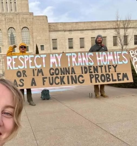 A group of protesters holding a large sign that reads "RESPECT MY TRANS HOMIES OR I'M GONNA IDENTIFY AS A FUCKING PROBLEM." The photo includes a person in the foreground, likely taking a selfie, while the protesters stand back behind the sign.