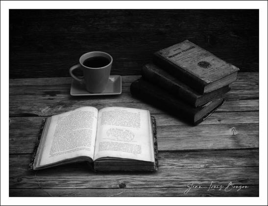 A grayscale still life composition.
A stack of three old books rests on a wooden table. The books have aged, darkened spines and appear to be thick volumes with text on the visible pages.
An open book, also appearing old, lies alongside the stack of books, lying open horizontally on the surface of the table and exposing the pages' text.
A cup of coffee sits alongside a saucer.
The wooden table has a rough-hewn, weathered texture. The wood grain is moderately visible.
The lighting in the image is soft and diffused, casting minimal shadows, highlighting the volumes and the cup of coffee. 