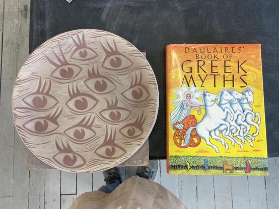 Carved platter with expressive eyes, book of Greek myths on a table 